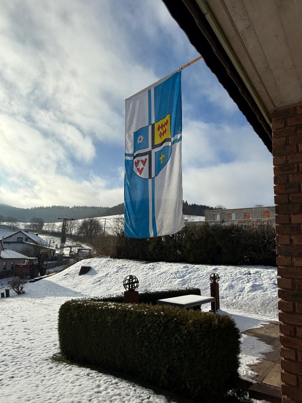 Eine vertikale Flagge mit dem Wappen von Kaltenborn, die an einem sonnigen Wintertag vor einem Haus und einer schneebedeckten Hügellandschaft weht.