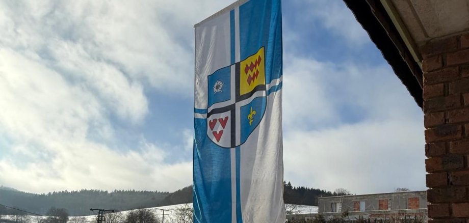 Eine vertikale Flagge mit dem Wappen von Kaltenborn, die an einem sonnigen Wintertag vor einem Haus und einer schneebedeckten Hügellandschaft weht.