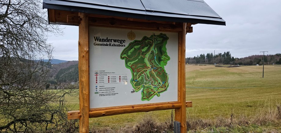 Ein Foto, zeigt eine neue, überdachte Informationstafel aus Holz mit einer großen Wanderwegekarte für die Gemeinde Kaltenborn. Die Tafel steht am Rand einer Wiese. Die Karte ist detailliert und farbig, mit verschiedenen markierten Wanderrouten in Grün- und Brauntönen. Links von der Karte befindet sich eine Legende mit Symbolen und kurzen Texten. Im Hintergrund erstreckt sich eine hügelige Landschaft mit bewaldeten Hängen. Der Himmel ist bedeckt und grau, was auf bewölktes Wetter hinweist.