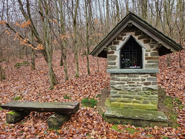 Ein kleiner, aus Natursteinen gemauerter Bildstock mit schiefergedecktem Satteldach steht am Rand eines bewaldeten Hangs. Der Bildstock hat eine spitzbogenförmige Öffnung mit einem kunstvoll geschmiedeten Eisengitter, durch das man einen kleinen Altar mit einer Figur im Inneren erkennen kann. Der Sockel des Bauwerks ist leicht moosbewachsen. Links neben dem Bildstock steht eine einfache, rustikale Bank, die aus einem dicken aufgetrennten Holzstamm als Sitzfläche und zwei quer liegenden Baumstämmen als Beinen besteht. Der Boden der gesamten Szene ist dicht mit braunem Herbstlaub bedeckt. Die umgebenden Bäume sind kahl, was auf die Jahreszeit Herbst oder Winter hinweist. Der Himmel ist bedeckt und grau.