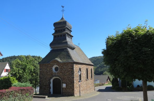 Kleine historische Steinkirche mit steilem Dach und mehrstufigem Glockenturm. Über dem Eingang ein Rundfenster, seitlich ein Bogenfenster. Umgeben von Bäumen, Büschen und weiteren Gebäuden unter blauem Himmel.