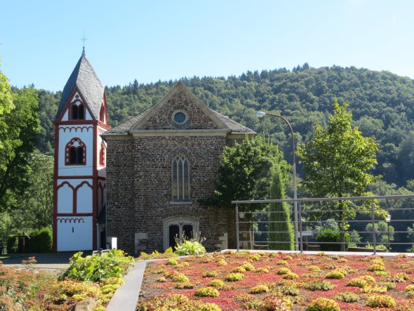Kirche mit weißer Fassade, roten Akzenten und spitzem Turm, daneben ein steinernes Gebäude. Davor gepflegter Garten mit Blumen, im Hintergrund bewaldeter Hügel unter blauem Himmel.
