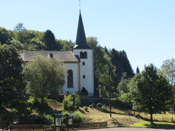 Kirche mit weißer Fassade, braunen Fensterrahmen und hohem Turm auf einem kleinen Hügel, umgeben von Bäumen. Vor der Kirche ein Zaun und zwei Infotafeln, im Hintergrund dichter Wald unter blauem Himmel.