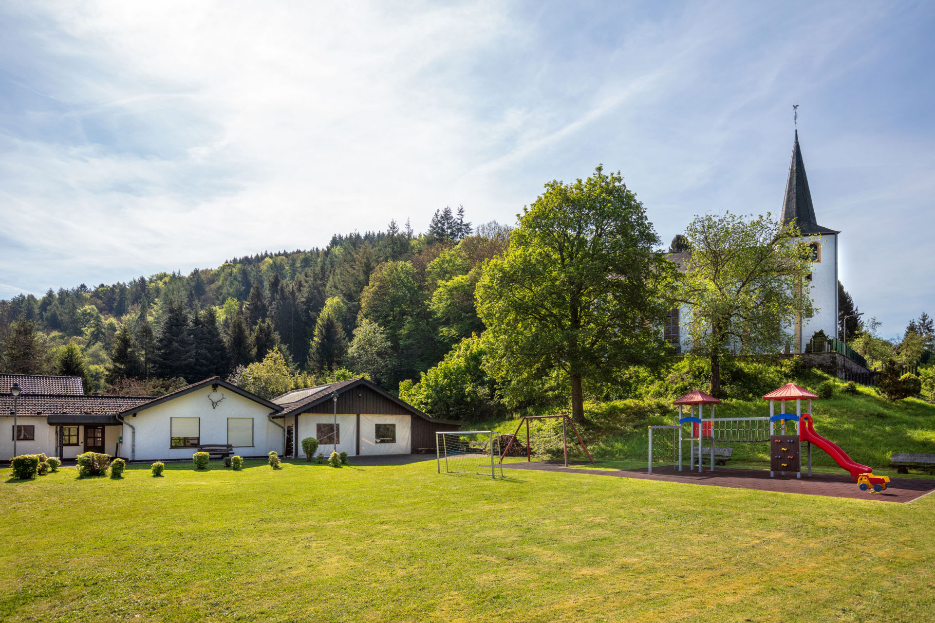 Grüne Wiese mit Spielplatz in der Mitte, ausgestattet mit bunten Rutschen und Klettergerüsten. Links zwei eingeschossige weiße Gebäude, rechts ein großer Baum. Im Hintergrund bewaldeter Hügel mit weißer Kirche und Turm unter blauem Himmel.