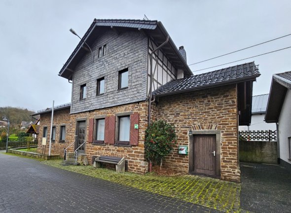 Ein Foto zeigt ein historisch wirkendes Gebäudeensemble, das aus einem zweistöckigen, schieferverkleideten Teil und einem einstöckigen, aus Natursteinen gemauerten Anbau besteht. Das Gebäude verfügt über eine dunkle Holztür im Steinanbau und eine weitere in der Schieferfassade. Fenster mit braunen Holzrahmen und teilweise roten Fensterläden sind zu sehen. Vor dem Gebäude verläuft ein Gehweg aus dunklem Pflasterstein. Eine einfache Holzbank steht vor einem Fenster des Steinanbaus. Ein kleiner, immergrüner Strauch mit roten Beeren wächst an der Ecke der beiden Gebäudeteile. Im Hintergrund sind weitere Wohnhäuser und ein bewaldeter Hügel unter einem bedeckten, grauen Himmel zu erkennen.