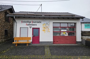 Ein Foto zeigt das Gebäude der Freiwilligen Feuerwehr Kaltenborn, ein weiß verputztes Gebäude mit Satteldach und einer roten Garagentor sowie einer roten Eingangstür. Neben der Eingangstür befindet sich ein kleines Fenster mit einer weißen Jalousie. An der Wand, zwischen der Tür und dem Garagentor, ist ein Wandgemälde, das den heiligen Florian darstellt. Über dem Gemälde steht in roter, geschwungener Schrift: „Freiwillige Feuerwehr Kaltenborn“. Vor dem Gebäude steht eine Holzbank. Der Vorplatz ist mit dunklen, quadratischen Pflastersteinen ausgelegt. Links vom Feuerwehrhaus grenzt ein älteres Gebäude mit einer Natursteinfassade und Schieferdach an. Im Hintergrund ist ein grauer, bedeckter Himmel zu sehen, was auf bewölktes Wetter hinweist.