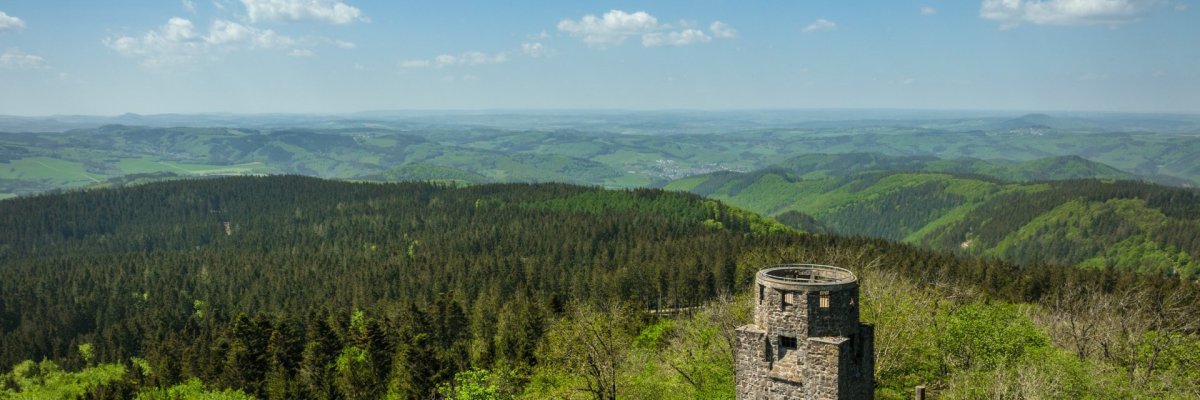 Steinerner Turm inmitten eines grünen Waldes, umgeben von Hügeln und Tälern. Der Himmel ist klar mit wenigen Wolken.