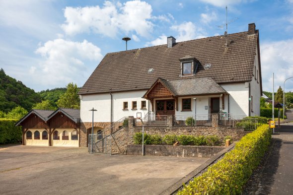 Zweistöckiges weißes Haus mit grauem Dach und breiter Treppe, daneben ein Nebengebäude mit Holzwand und Toren, umgeben von Asphaltfläche und grüner Hügellandschaft.