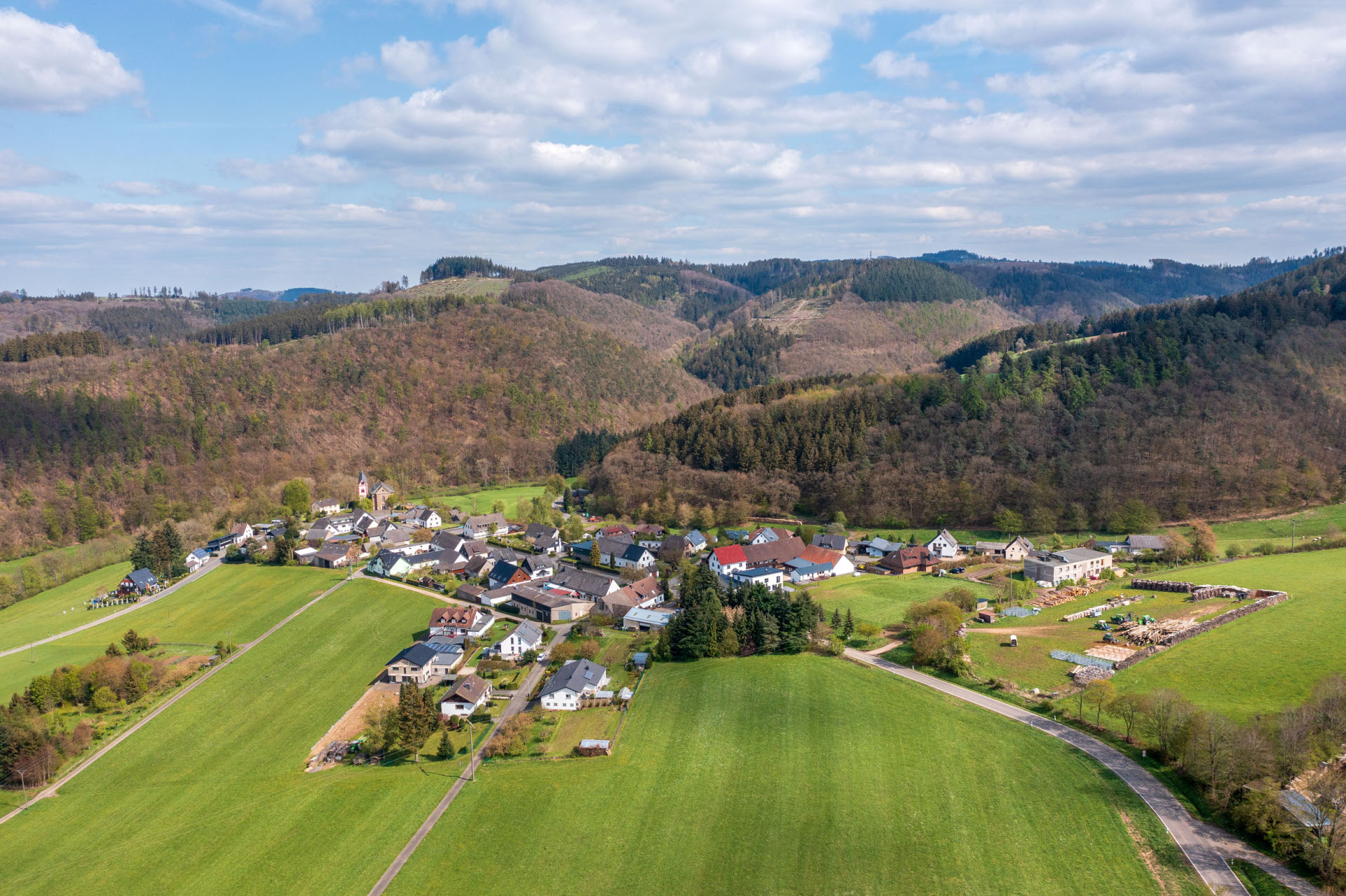 Luftaufnahme Ort Kaltenborn mit Häusern und Straßen, umgeben von grünen Feldern und bewaldeten Hügeln. Der blaue Himmel ist teils bewölkt.