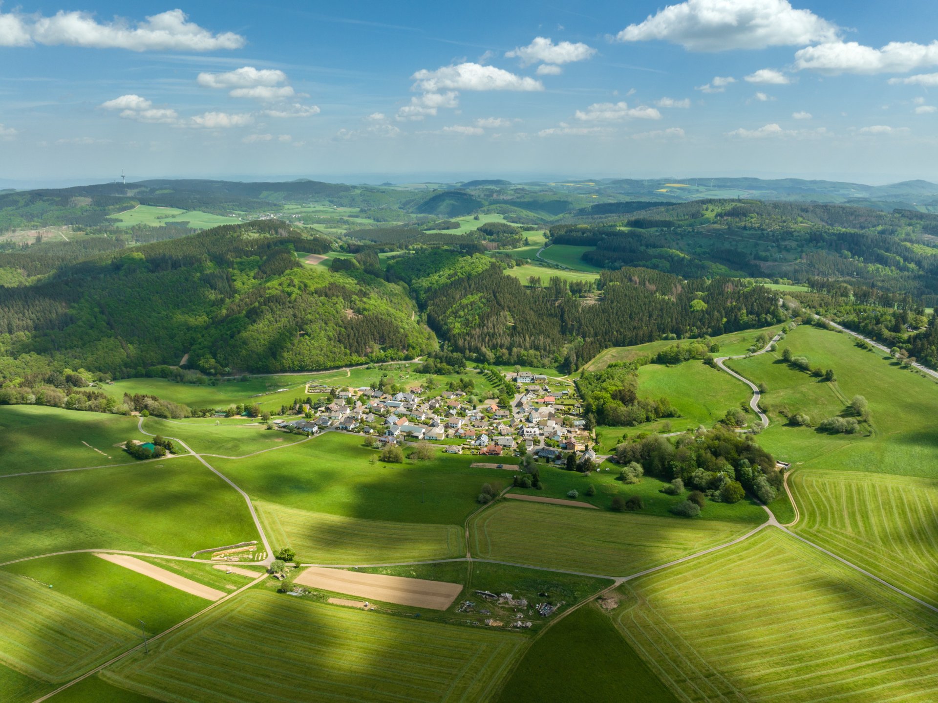 Luftaufnahme Ortsteil Jammelshofen in einem Tal, umgeben von grünen Feldern, Wiesen und dichten Wäldern. Im Hintergrund sanfte Hügel, darüber ein Himmel mit vereinzelten Wolken.