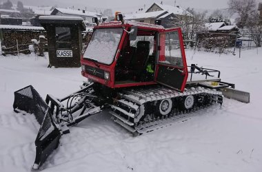 Rotes Pistenfahrzeug im Schnee vor einem kleinen Gebäude mit der Aufschrift ‚KASSE‘. Im Hintergrund schneebedeckte Häuser und Bäume.