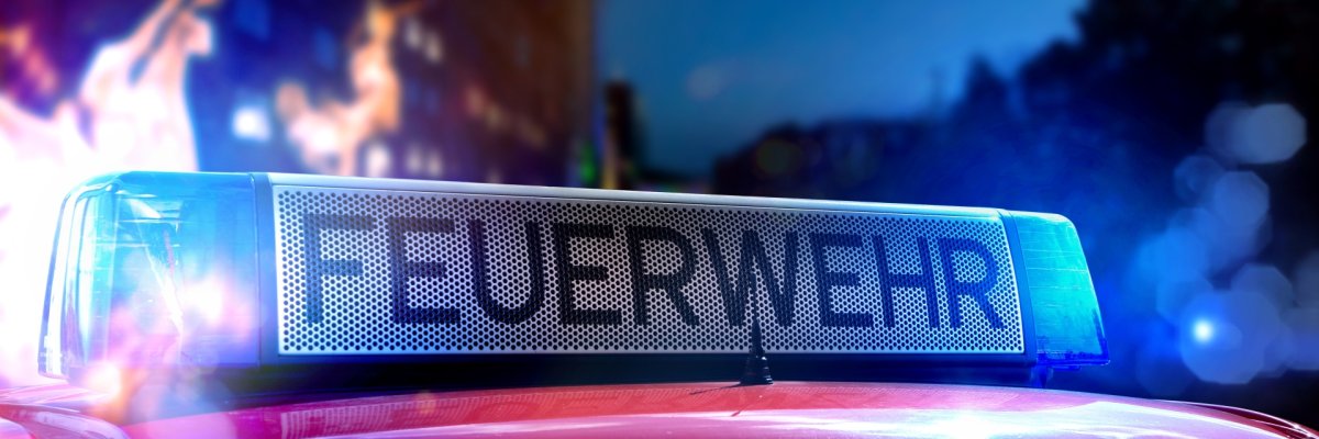 Ein Feuerwehrfahrzeug mit leuchtendem 'FEUERWEHR'-Schild auf dem Dach. Im Hintergrund sind Flammen und Lichter zu erkennen – es wirkt wie ein Einsatzort bei Nacht.