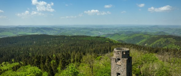 Steinerner Turm inmitten eines grünen Waldes, umgeben von Hügeln und Tälern. Der Himmel ist klar mit wenigen Wolken.
