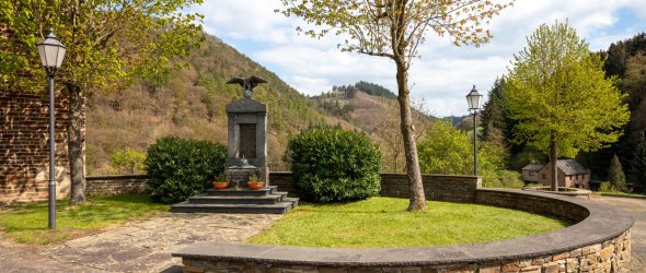 Ein gepflegter Gedenkort mit einem steinernen Denkmal und Adlerfigur, umgeben von Pflanzen, einem Rundweg und grüner Hügellandschaft im Hintergrund.