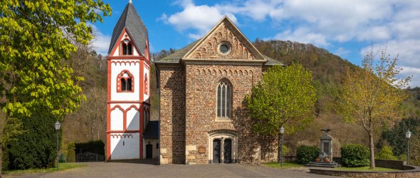 Kirche in Kaltenborn mit weißem Turm und rotem Zierwerk links, steinernem Kirchenschiff mit Rundbogenfenstern rechts. Umgeben von Bäumen, davor gepflasterter Platz, im Hintergrund bewaldete Hügel unter teils bewölktem blauem Himmel.
