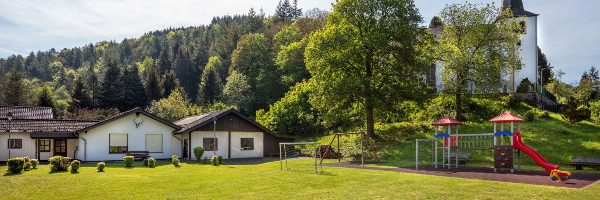 Grüne Wiese mit Spielplatz in der Mitte, ausgestattet mit bunten Rutschen und Klettergerüsten. Links zwei eingeschossige weiße Gebäude, rechts ein großer Baum. Im Hintergrund bewaldeter Hügel mit weißer Kirche und Turm unter blauem Himmel.
