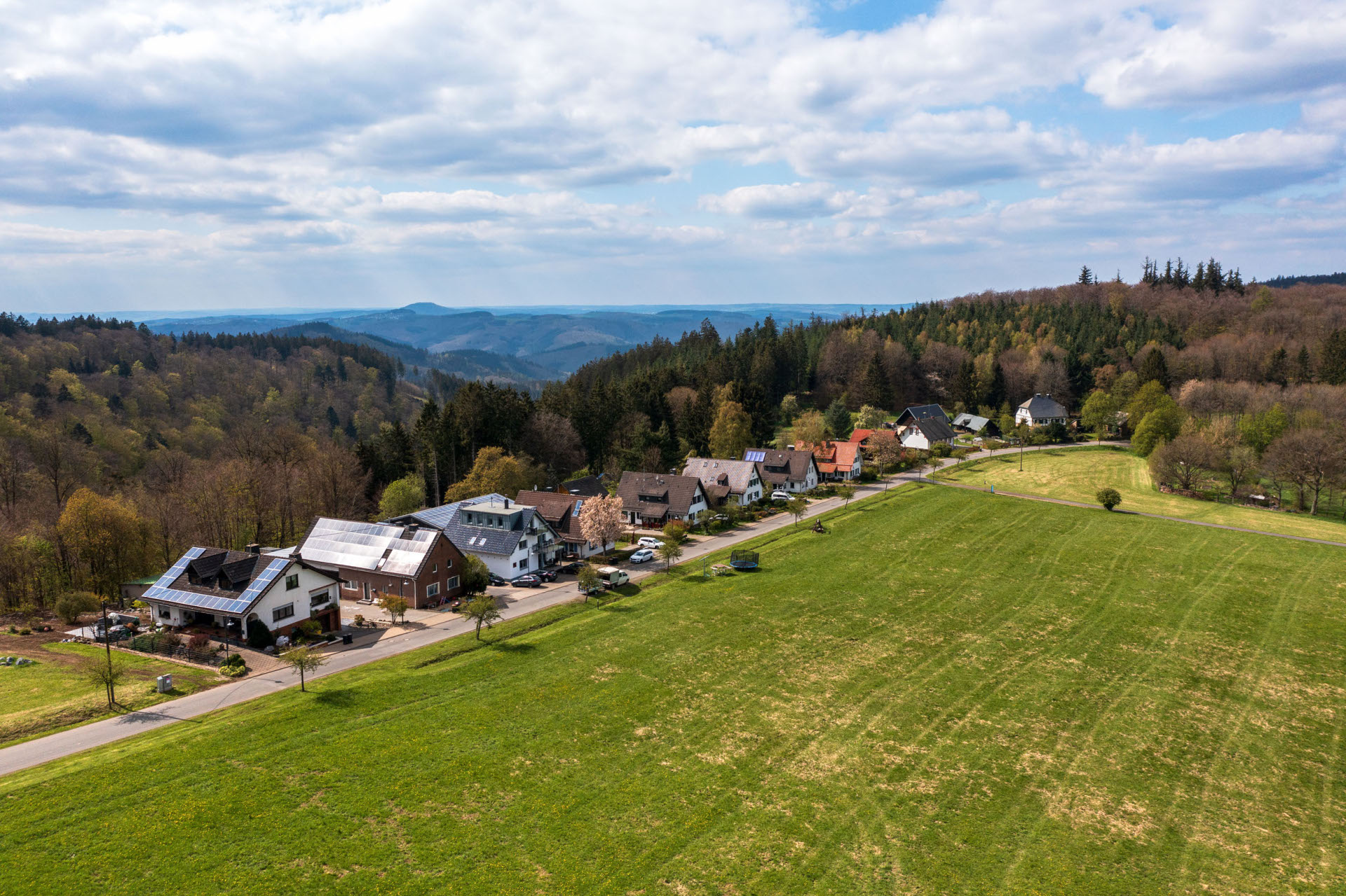 Aufnahme aus der Vogelperspektive auf den Ort Hochacht mit mehreren Häusern entlang einer Straße, umgeben von Wiesen und Feldern. Im Hintergrund bewaldete Hügel unter teils bewölktem Himmel.