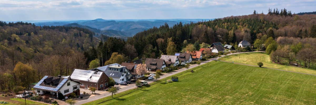 Aufnahme aus der Vogelperspektive auf den Ort Hochacht mit mehreren Häusern entlang einer Straße, umgeben von Wiesen und Feldern. Im Hintergrund bewaldete Hügel unter teils bewölktem Himmel.