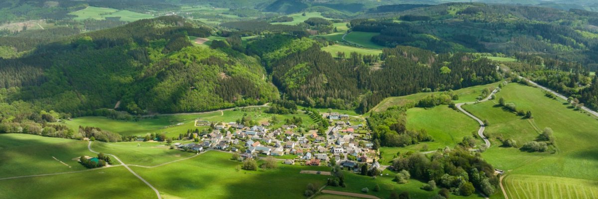Luftaufnahme Ortsteil Jammelshofen in einem Tal, umgeben von grünen Feldern, Wiesen und dichten Wäldern. Im Hintergrund sanfte Hügel, darüber ein Himmel mit vereinzelten Wolken.