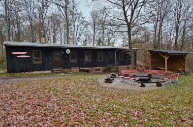 Ein Foto zeigt eine langgestreckte, einstöckige Holzhütte mit dunkel gestrichener Fassade und einem Blechdach, umgeben von einem Wald und einer Wiese. Die Hütte wirkt rustikal und verfügt über mehrere kleine Fenster, eine braune Eingangstür in der Mitte und drei Schornsteine auf dem Dach. Auf der rechten Seite des Gebäudes gibt es einen kleinen, offenen Unterstand. Im Vordergrund befindet sich ein befestigter Grill- oder Lagerfeuerplatz, um den mehrere rote Bänke mit Metallgestellen gruppiert sind. Der Boden der Wiese ist mit feuchtem, braunem Herbstlaub bedeckt. Die umgebenden Bäume sind kahl, und der Himmel ist gleichmäßig bedeckt und grau, was auf einen kühlen, bewölkten Tag hinweist.