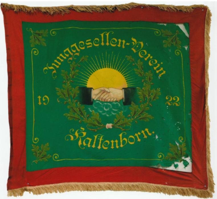 Quadratische Vereinsfahne mit rotem Rand und goldenen Fransen. In der Mitte ist ein grünes Feld mit gelber Schrift. Darauf steht: „Junggesellen-Verein Kaltenborn 1922“. Im Zentrum der Fahne sind zwei Hände dargestellt, die sich zum Handschlag reichen. Hinter den Händen ist eine große gelbe Sonne mit Strahlen zu sehen. Um die Hände herum sind grüne Eichenblätter angeordnet.