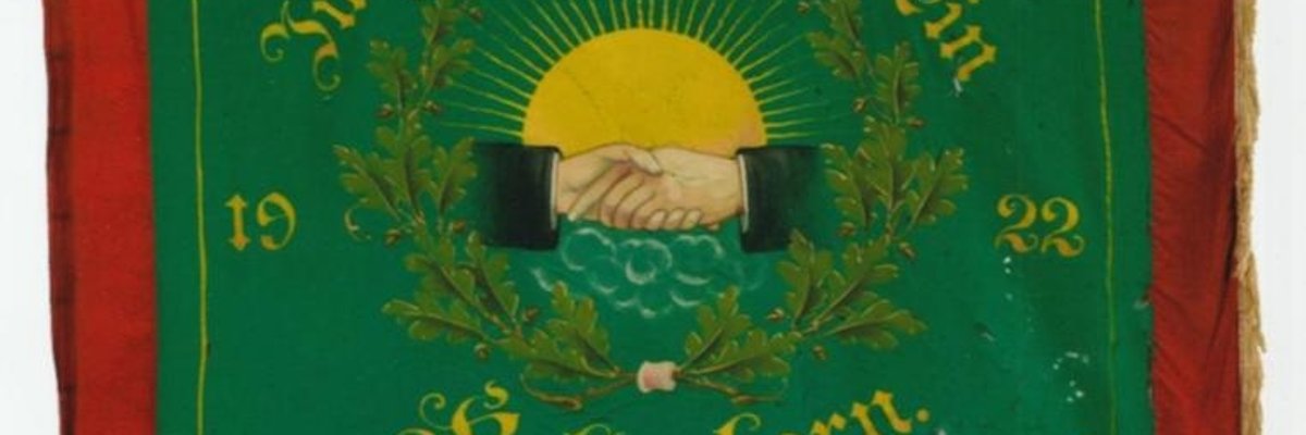 Quadratische Vereinsfahne mit rotem Rand und goldenen Fransen. In der Mitte ist ein grünes Feld mit gelber Schrift. Darauf steht: „Junggesellen-Verein Kaltenborn 1922“. Im Zentrum der Fahne sind zwei Hände dargestellt, die sich zum Handschlag reichen. Hinter den Händen ist eine große gelbe Sonne mit Strahlen zu sehen. Um die Hände herum sind grüne Eichenblätter angeordnet.