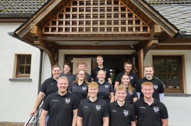 Eine Gruppe von Personen steht auf einer Treppe vor einem Gebäude mit weißer Fassade und einem Holzdach über dem Eingang. Das Dach hat ein dekoratives Holzgitter. Die Personen tragen schwarze Poloshirts mit einem weißen Vereinslogo  JGV Kaltenborn, auf der linken Brustseite. Einige Shirts haben zusätzlich Namen aufgedruckt.