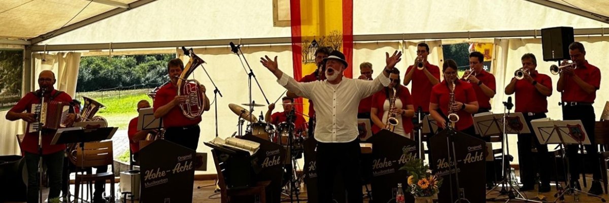 Blaskapelle in roter Kleidung spielt unter einem großen Festzelt, mit Instrumenten wie Trompeten, Akkordeon und Schlagzeug. Im Vordergrund steht ein Dirigent mit erhobenen Armen, hinter ihm eine gelb-rote Fahne