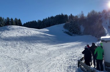 Eine Aufnahme einer sonnigen, verschneiten Skipiste mit mehreren Skifahrern oder Snowboardern am unteren rechten Rand, die vor einem kleinen Gebäude stehen. Der Hang ist mit Schnee bedeckt und weist viele Spuren auf. Im Hintergrund säumen schneebedeckte Tannen den Hügel, und die Sonne scheint hell am oberen rechten Rand, wo auch ein Skilift zu sehen ist.