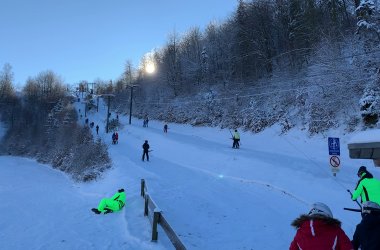Eine weitläufige Aufnahme einer Skipiste an einem sonnigen, verschneiten Tag, auf der mehrere Skifahrer und Snowboarder auf und neben dem Hang zu sehen sind, darunter eine Person in leuchtend neongrüner Kleidung, die auf dem Schnee sitzt, und eine andere Gruppe in der unteren rechten Ecke. Im Hintergrund führt ein Skilift den schneebedeckten Hang hinauf, gesäumt von schneebedeckten Bäumen, während die Sonne hell am oberen Rand des Bildes scheint.