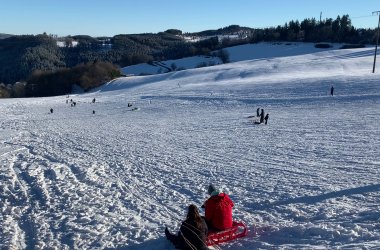 Eine Aufnahme eines schneebedeckten Rodelhangs an einem sonnigen Tag. Mehrere Menschen, hauptsächlich Kinder und Familien, sind auf dem Hang verteilt und rodeln oder spielen im Schnee. Im Vordergrund sitzen zwei Personen in Winterkleidung (eine trägt eine rote Jacke) auf einem roten Schlitten und blicken vom Betrachter weg. Im Hintergrund sind sanfte Hügel zu sehen, die mit Schnee und dunklen Nadelwäldern bedeckt sind, unter einem klaren blauen Himmel.