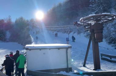 Eine  Aufnahme eines sonnigen, verschneiten Skihangs. Im Vordergrund, links von einem weißen Gebäude, stehen mehrere Skifahrer in Winterkleidung, darunter eine Person mit einer neongrünen Jacke. Rechts davon befindet sich der markante, obere Teil eines Schlepplifts. Der Hang erstreckt sich den Hügel hinauf, wo weitere Skifahrer und Snowboarder auf der Piste oder am Rand in der Nähe eines Liftmasts zu sehen sind. Der Hang ist von schneebedeckten Bäumen gesäumt, und die Sonne scheint hell am oberen linken Rand des Bildes, wodurch das Licht blendet.