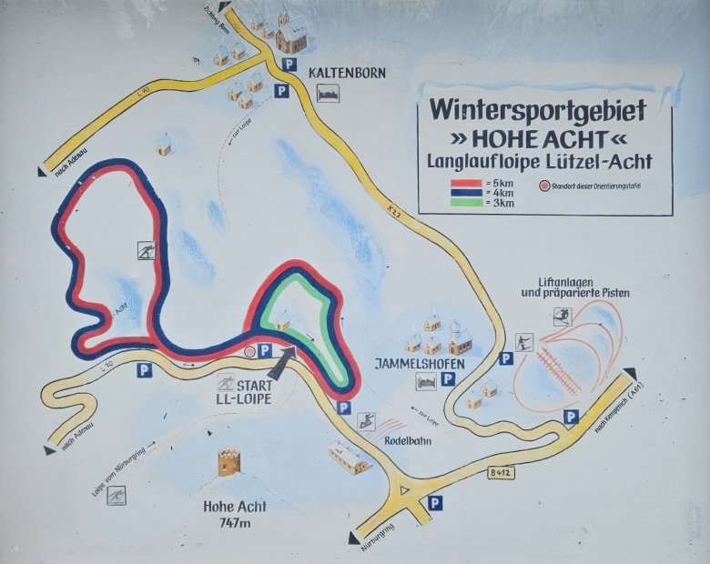 Eine farbige Karte zeigt das Wintersportgebiet Hohe Acht/Jammelshofen mit markierten Langlaufloipen und Pisten in der Eifel. Die Karte stellt das Gebiet topografisch dar, mit Straßen wie der K 23 und B 412, Ortschaften wie Kaltenborn und Jammelshofen sowie Parkplätzen (P). Eine Legende in der oberen rechten Ecke erklärt die farbliche Kennzeichnung der Langlaufloipen: Rot steht für 5 km, Blau für 4 km und Grün für 3 km. Es gibt auch Symbole für Liftanlagen, präparierte Pisten, eine Rodelbahn und den Standort der Orientierungstafel. Die Hohe Acht mit 747 Metern Höhe ist ebenfalls eingezeichnet. Der Hintergrund der Karte ist überwiegend weiß mit blauen Flächen, die Schneegebiete oder Wasser darstellen könnten.